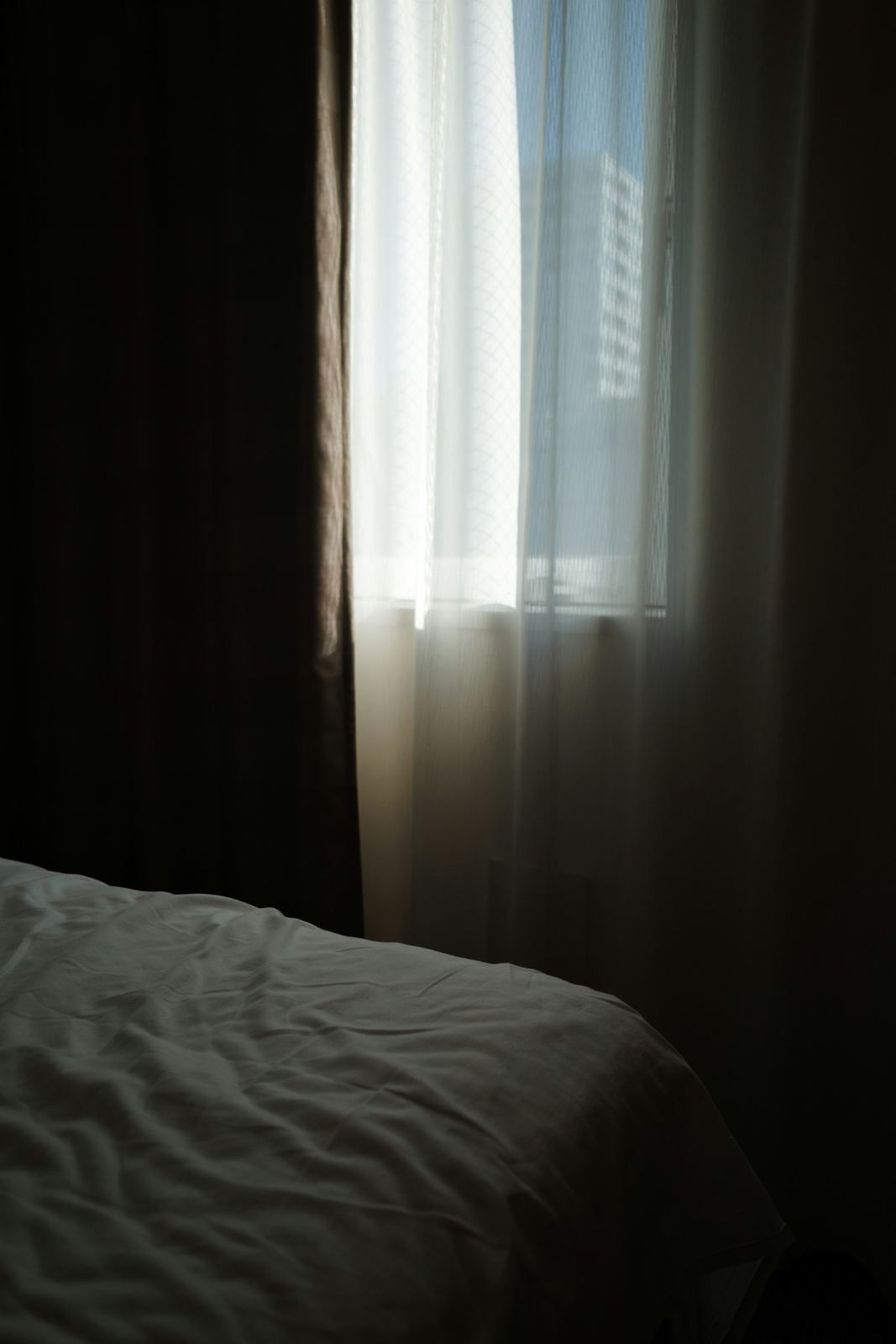 A dim bedroom with an unmade bed, bright daylight pouring through sheer curtains on one side of the room