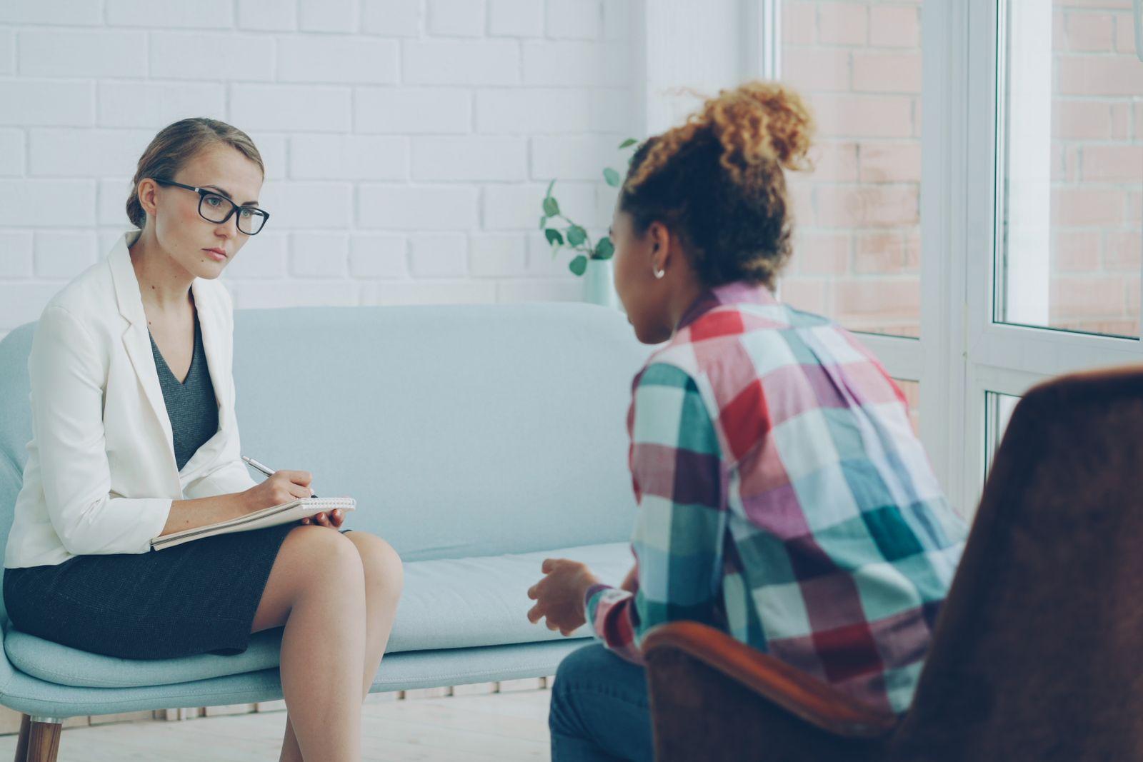 A therapist listening and taking notes while a client talks, in a bright room with plants